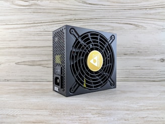 A computer power supply unit with a black casing, featuring a prominent circular fan grille. The center of the fan has a logo with a yellow background. The sides have a honeycomb grill, and there are visible power connectors, including an on/off switch. The unit sits on a light wooden surface with a textured wall in the background.