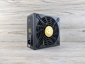A computer power supply unit with a black casing, featuring a prominent circular fan grille. The center of the fan has a logo with a yellow background. The sides have a honeycomb grill, and there are visible power connectors, including an on/off switch. The unit sits on a light wooden surface with a textured wall in the background.