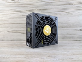 A computer power supply unit with a black casing, featuring a prominent circular fan grille. The center of the fan has a logo with a yellow background. The sides have a honeycomb grill, and there are visible power connectors, including an on/off switch. The unit sits on a light wooden surface with a textured wall in the background.