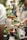 A group of people engaged in a gardening activity, with various potted plants and gardening tools arranged on a table. One person is tending to a plant in a terracotta pot, while others are interacting or observing in a greenery-filled space.