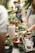 A group of people engaged in a gardening activity, with various potted plants and gardening tools arranged on a table. One person is tending to a plant in a terracotta pot, while others are interacting or observing in a greenery-filled space.