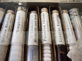 A collection of vintage medicine or chemical tubes arranged in a box. Each tube has a label with text indicating the substance inside, such as quinine bisulphate and bismuth and soda. The tubes appear to be made of glass and have cork stoppers, with some aging visible on the labels and corks.