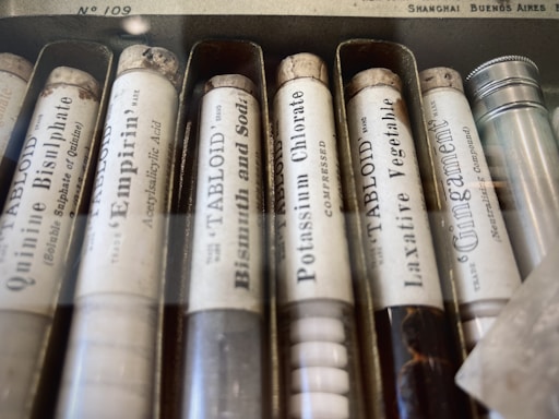 A collection of vintage medicine or chemical tubes arranged in a box. Each tube has a label with text indicating the substance inside, such as quinine bisulphate and bismuth and soda. The tubes appear to be made of glass and have cork stoppers, with some aging visible on the labels and corks.