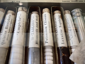A collection of vintage medicine or chemical tubes arranged in a box. Each tube has a label with text indicating the substance inside, such as quinine bisulphate and bismuth and soda. The tubes appear to be made of glass and have cork stoppers, with some aging visible on the labels and corks.