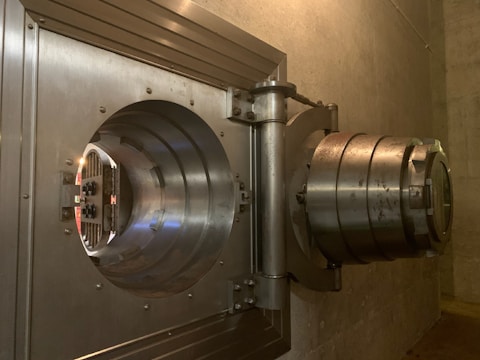 A large metal vault door, partially open, revealing a thick, reinforced structure. The door has several circular metal layers and appears to be located in a concrete room with a dim light.