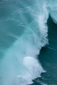 Powerful ocean waves crashing and creating a frothy white crest against a deep teal sea. The image shows the dynamic motion of water with a range of lighter and darker shades, giving a sense of raw natural energy.