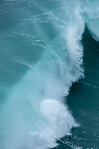 Powerful ocean waves crashing and creating a frothy white crest against a deep teal sea. The image shows the dynamic motion of water with a range of lighter and darker shades, giving a sense of raw natural energy.