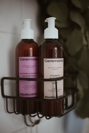 Two pump bottles of hair care products are placed in a black wire shower caddy. The bottle on the left has a purple label and contains conditioner, while the bottle on the right has a cream label and contains shampoo. Both bottles are branded 'Ceremonia' and have a minimalist design. The background is slightly blurred, featuring leaves.
