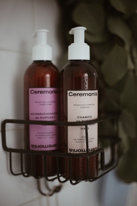 Two pump bottles of hair care products are placed in a black wire shower caddy. The bottle on the left has a purple label and contains conditioner, while the bottle on the right has a cream label and contains shampoo. Both bottles are branded 'Ceremonia' and have a minimalist design. The background is slightly blurred, featuring leaves.
