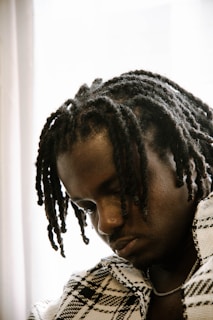 A person with medium-length, textured dreadlocks gazes downward with a contemplative expression. The individual is wearing a patterned jacket, and the soft lighting creates a warm, introspective atmosphere.
