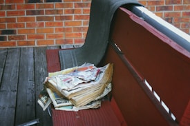 A bundle of old, yellowed newspapers sits on a wooden bench. The bench is painted red, and a fabric material is draped over the backrest. The background features a brick wall and a worn wooden floor, suggesting an outdoor or porch setting.