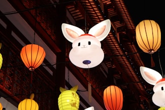 Luminous rabbit-shaped lanterns and traditional round lanterns hang against the backdrop of an ornate, wooden building facade, richly decorated in red and intricate designs.