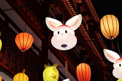 Luminous rabbit-shaped lanterns and traditional round lanterns hang against the backdrop of an ornate, wooden building facade, richly decorated in red and intricate designs.