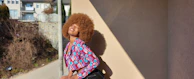 A person with a large afro hairstyle is smiling and leaning back against a beige wall, casting a shadow. The individual is wearing a colorful, patterned jacket. In the background, there are buildings and some bare shrubs along a path.