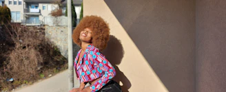 A person with a large afro hairstyle is smiling and leaning back against a beige wall, casting a shadow. The individual is wearing a colorful, patterned jacket. In the background, there are buildings and some bare shrubs along a path.