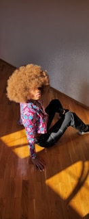 A person with a large, curly afro hairstyle is sitting on a wooden floor. They are wearing a colorful patterned shirt and dark pants. Sunlight casts shadows on the floor, giving a warm and vibrant atmosphere.
