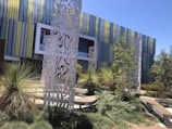A modern architectural building with a facade of vertical colored panels in shades of blue and green. In front of the building are intricately designed metal sculptures surrounded by landscaped greenery, including various bushes and small trees. The scene is well-lit by sunlight, casting shadows on the ground.