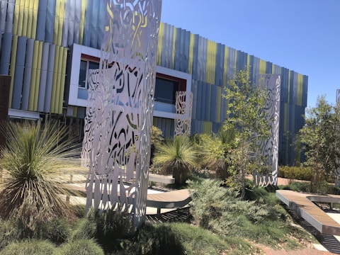 A modern architectural building with a facade of vertical colored panels in shades of blue and green. In front of the building are intricately designed metal sculptures surrounded by landscaped greenery, including various bushes and small trees. The scene is well-lit by sunlight, casting shadows on the ground.