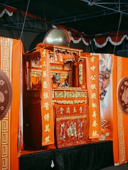 A vibrant and ornate red shrine with intricate gold detailing, decorated with vertical orange banners featuring Chinese calligraphy. The shrine is adorned with traditional symbols and motifs, and there is a round metallic dome on top. The background includes a partially visible, elaborately painted dragon design.