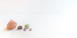 Five gemstones of varying shapes and colors are placed on a light, neutral background. The stones include a large pinkish-orange crystal, a shiny metallic stone, a light green stone, a small purple stone, and a smooth, pale pink pebble.