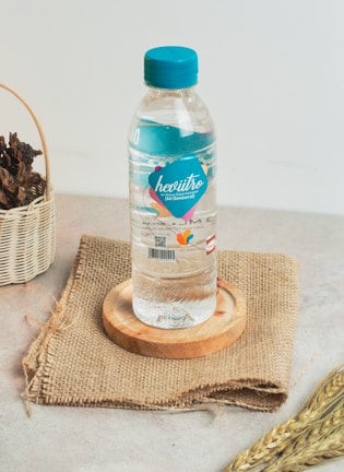 A plastic bottle of mineral water with a blue cap is placed on a wooden coaster, which is on top of a piece of burlap cloth. To the left, there's a wicker basket containing dried leaves. In the foreground, a few stalks of wheat are visible.