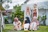 A group of five people wearing hijabs are gathered outdoors in a garden setting. Three of them are sitting on the grass while two are standing. They appear to be engaged in a conversation and are smiling. In the background, there is a white ornate building and some green plants.