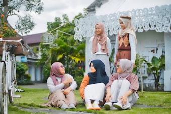 A group of five people wearing hijabs are gathered outdoors in a garden setting. Three of them are sitting on the grass while two are standing. They appear to be engaged in a conversation and are smiling. In the background, there is a white ornate building and some green plants.