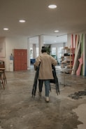 A person dressed in a beige coat and jeans is walking away from the camera, carrying several chairs. The setting appears to be an indoor workspace or studio with concrete floors, overhead lights, red lockers, a water dispenser, tables, and various rolled-up materials leaning against a wall. The space opens into a room with windows and natural light streaming in.