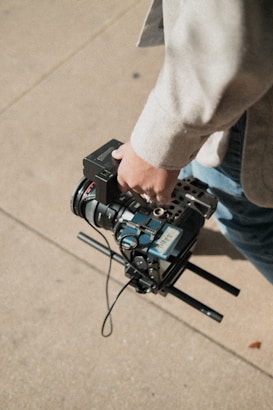 A person holding a professional video camera with various components including a lens, cables, and handles, while standing on a light-colored concrete surface.