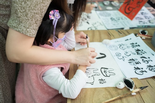 A child, assisted by an adult, is practicing calligraphy on white paper. The table is scattered with brushes, ink, and sheets of paper containing various drawings and Chinese characters. The child is dressed in a pink outfit and appears focused on the task.