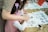 A child, assisted by an adult, is practicing calligraphy on white paper. The table is scattered with brushes, ink, and sheets of paper containing various drawings and Chinese characters. The child is dressed in a pink outfit and appears focused on the task.