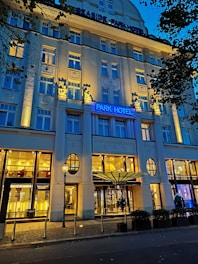 An elegant hotel facade lit by warm yellow lights against a deep blue evening sky. The building features decorative columns and large windows, with a prominent sign displaying 'Park Hotel.' Trees with dark leaves frame the image on one side.