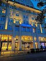 An elegant hotel facade lit by warm yellow lights against a deep blue evening sky. The building features decorative columns and large windows, with a prominent sign displaying 'Park Hotel.' Trees with dark leaves frame the image on one side.