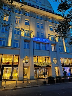 An elegant hotel facade lit by warm yellow lights against a deep blue evening sky. The building features decorative columns and large windows, with a prominent sign displaying 'Park Hotel.' Trees with dark leaves frame the image on one side.