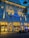 An elegant hotel facade lit by warm yellow lights against a deep blue evening sky. The building features decorative columns and large windows, with a prominent sign displaying 'Park Hotel.' Trees with dark leaves frame the image on one side.