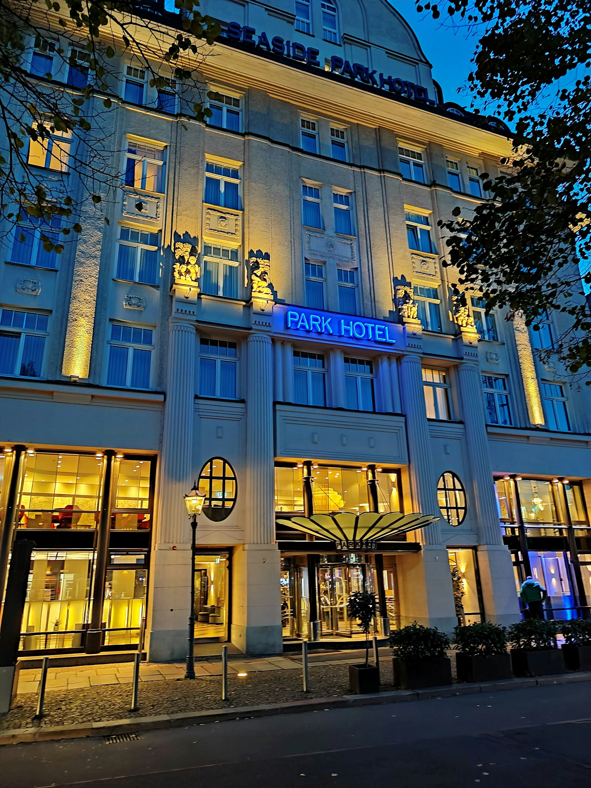An elegant hotel facade lit by warm yellow lights against a deep blue evening sky. The building features decorative columns and large windows, with a prominent sign displaying 'Park Hotel.' Trees with dark leaves frame the image on one side.