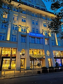 An elegant hotel facade lit by warm yellow lights against a deep blue evening sky. The building features decorative columns and large windows, with a prominent sign displaying 'Park Hotel.' Trees with dark leaves frame the image on one side.