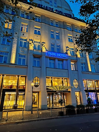 An elegant hotel facade lit by warm yellow lights against a deep blue evening sky. The building features decorative columns and large windows, with a prominent sign displaying 'Park Hotel.' Trees with dark leaves frame the image on one side.