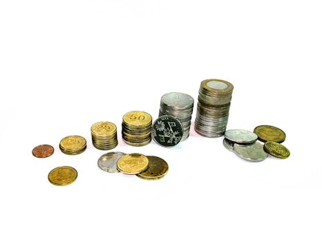 A collection of coins arranged in various stacks and scattered across a plain white background. Coins vary in size, color, and denomination, including some gold-colored and silver-colored ones, with visible inscriptions and designs.