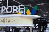 Neon and painted signs with blurred words and letters. Includes words like 'SANDWICH' and partial letters of another word. The backdrop features different-painted designs of letter W in green, blue, and yellow colors.