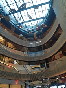 A modern architectural interior featuring multiple levels with metallic railings and a large glass ceiling. Suspended decorative elements hang from above, and various floors of retail stores are visible, including recognizable brand signs.