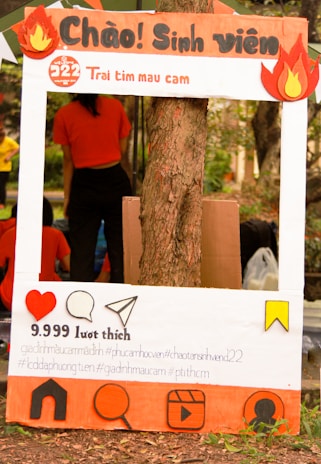 A decorated cardboard frame designed to look like a social media post is set up against a tree, featuring Vietnamese text and various social media icons. The frame includes cutouts of flames at the top corners and a large heart icon, speech bubble, paper airplane, and other symbols at the bottom. Behind the frame is a person in an orange shirt, with other people visible in the background.