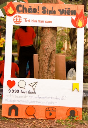 A decorated cardboard frame designed to look like a social media post is set up against a tree, featuring Vietnamese text and various social media icons. The frame includes cutouts of flames at the top corners and a large heart icon, speech bubble, paper airplane, and other symbols at the bottom. Behind the frame is a person in an orange shirt, with other people visible in the background.
