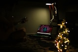 In a dimly lit room, a person plays an acoustic guitar next to an illuminated laptop displaying a futuristic interface. A small, decorated Christmas tree with glowing lights appears prominently in the foreground. A lamp above the chair provides a soft glow.