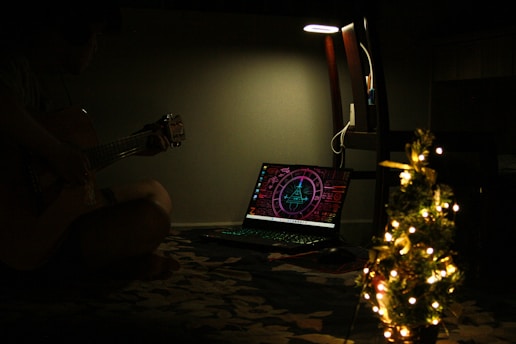 In a dimly lit room, a person plays an acoustic guitar next to an illuminated laptop displaying a futuristic interface. A small, decorated Christmas tree with glowing lights appears prominently in the foreground. A lamp above the chair provides a soft glow.