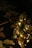A small, decorative Christmas tree adorned with lights and golden ornaments is placed against a dark background. The tree is illuminated, creating a warm, festive glow. A guitar neck is partially visible behind the tree, lying on a patterned fabric with floral motifs.