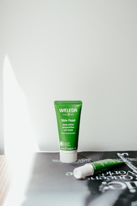 A green tube of Weleda Skin Food body lotion is standing upright on a surface, with a second similar tube lying on its side next to it. The background is neutral, and the tubes are positioned under soft lighting creating shadows.