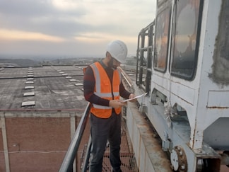 A person wearing a white hard hat and an orange safety vest is standing on a high platform, examining a piece of paper. The background shows a vast industrial area with a cloudy sky.