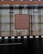 The image features a concrete wall with a decorative batik pattern in the center, set above a roadway with a black and white striped curb. There is text beneath the pattern, reading 'BATIK CEPLOK SEKAR KOPI.' Water stains are visible on the concrete surface, indicating possible weather exposure.