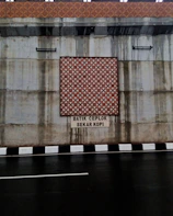 The image features a concrete wall with a decorative batik pattern in the center, set above a roadway with a black and white striped curb. There is text beneath the pattern, reading 'BATIK CEPLOK SEKAR KOPI.' Water stains are visible on the concrete surface, indicating possible weather exposure.